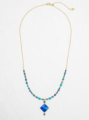 A gold chain necklace featuring a central blue diamond-shaped pendant and beaded strands of blue, teal, and dark gray beads, displayed on a textured white background.
