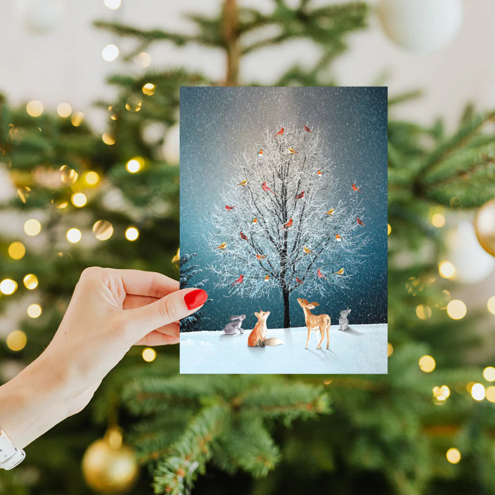 Hand holding a Christmas card with animals in front of a decorated tree.
