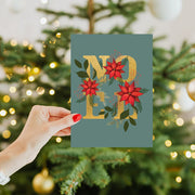 Hand holding a Christmas card with 'Merry Christmas' text in front of a decorated tree.