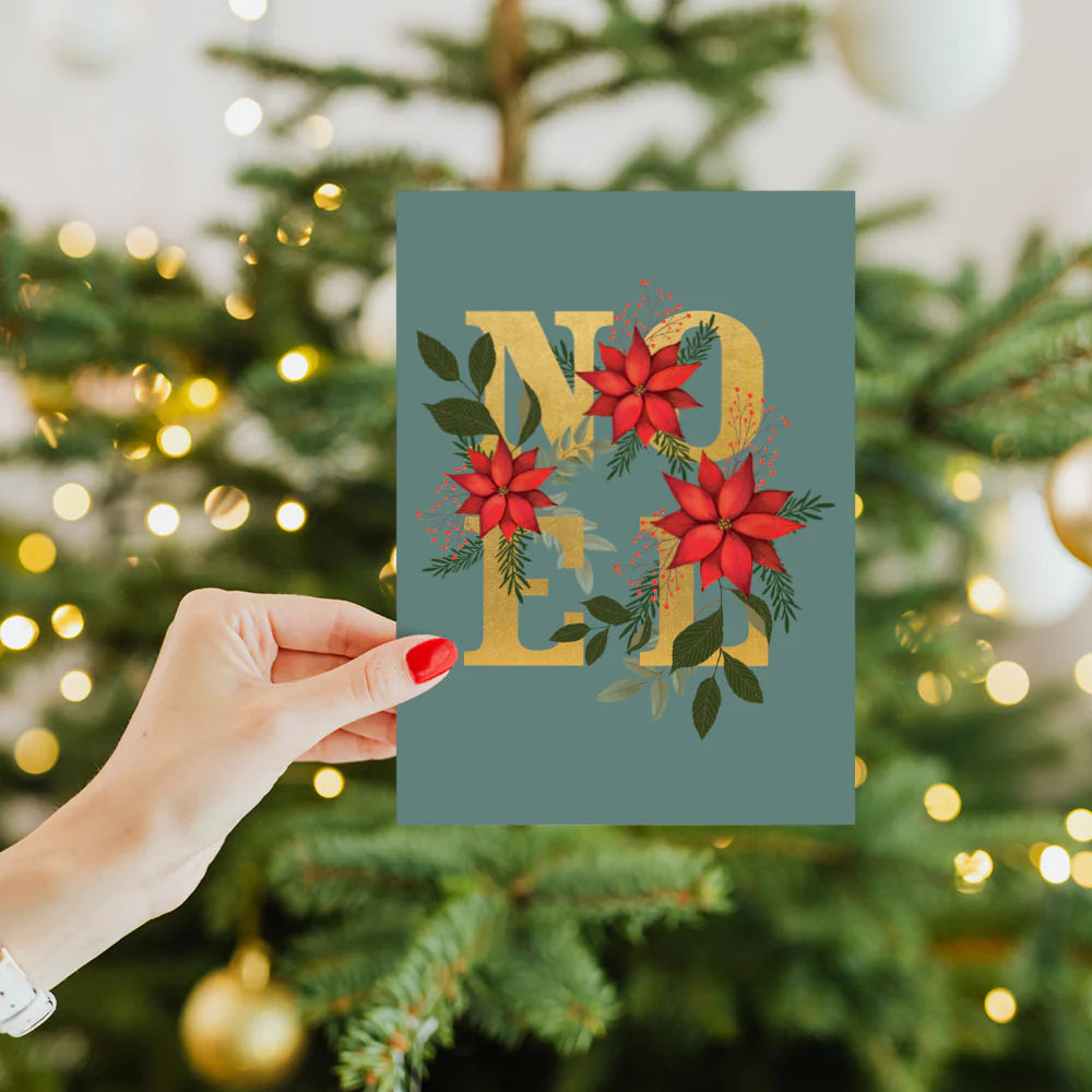 Hand holding a Christmas card with 'Merry Christmas' text in front of a decorated tree.