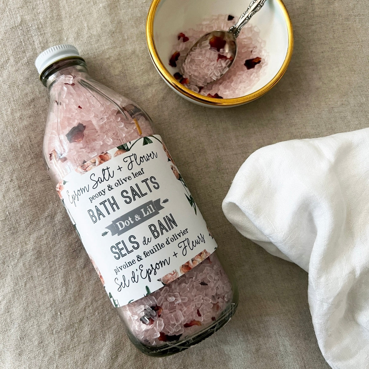 Bottle of bath salts with a label, bowl of bath salts, and towel on a textured surface