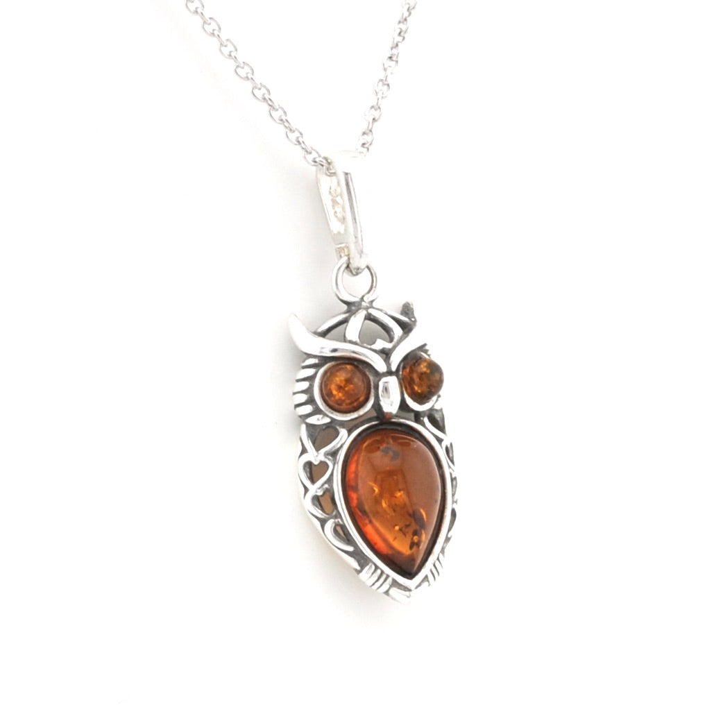 An owl pendant necklace featuring a silver-toned metal frame with a large teardrop-shaped amber gemstone in the center and two smaller round amber gemstones for the eyes, suspended from a silver chain on a white background.