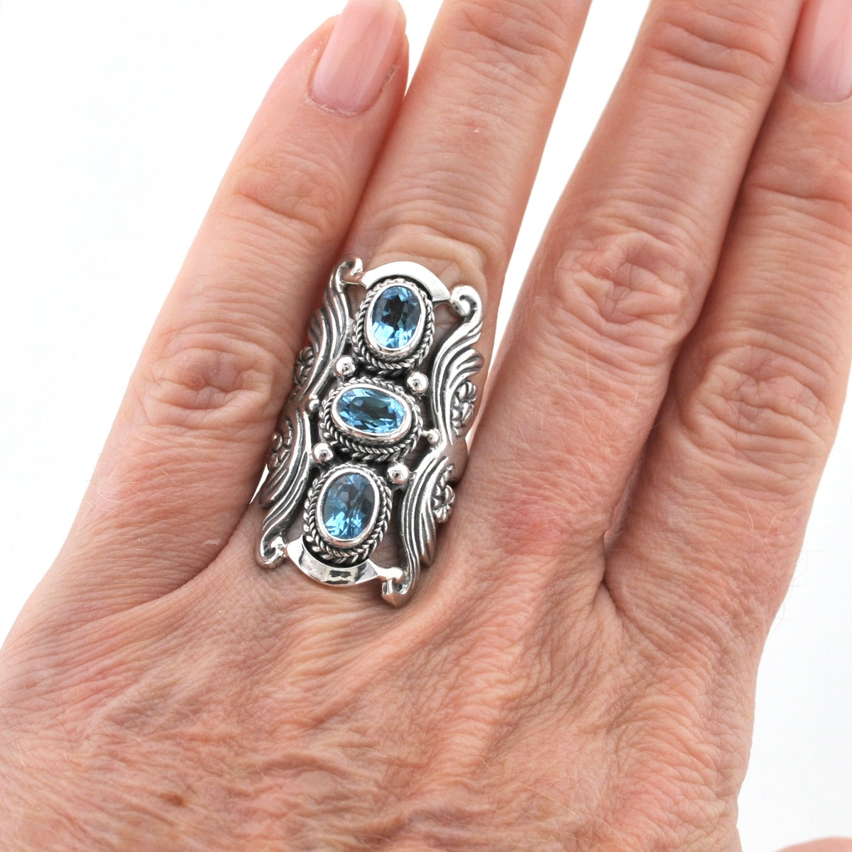 A close-up of a hand wearing a large silver ring with three oval light blue gemstones, set against a plain white background.