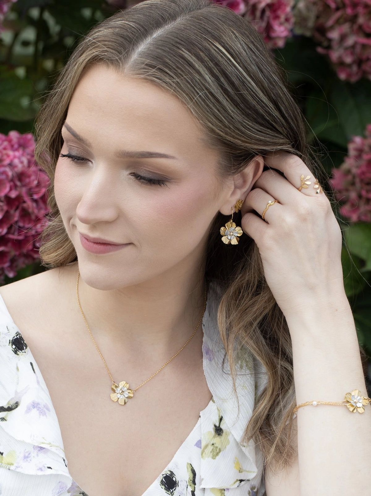 Woman wearing gold jewelry including earrings, necklace, and bracelet with a floral background