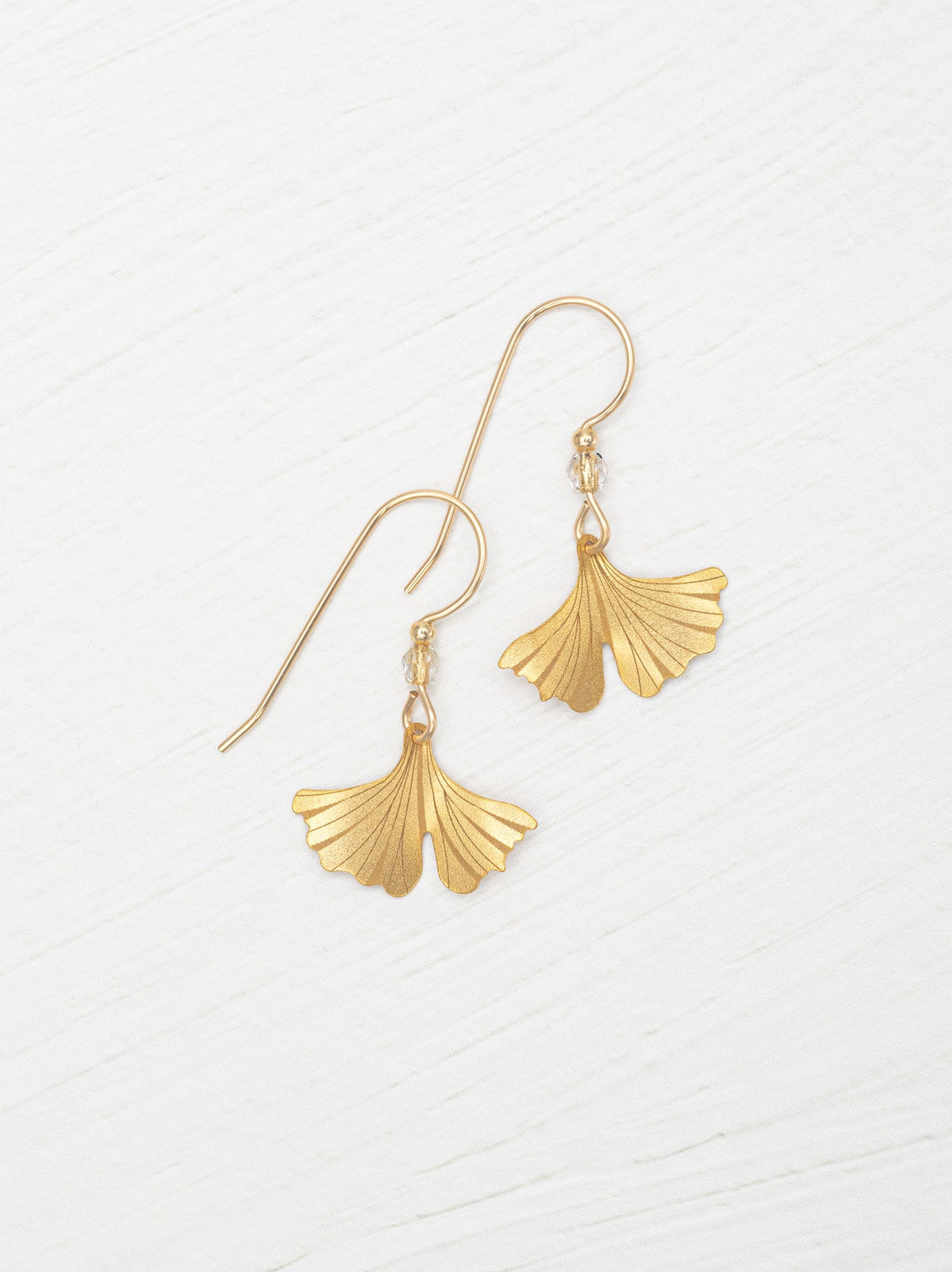 A pair of gold-tone dangle earrings with fan-shaped ginkgo leaf pendants, displayed on a light, textured white background.