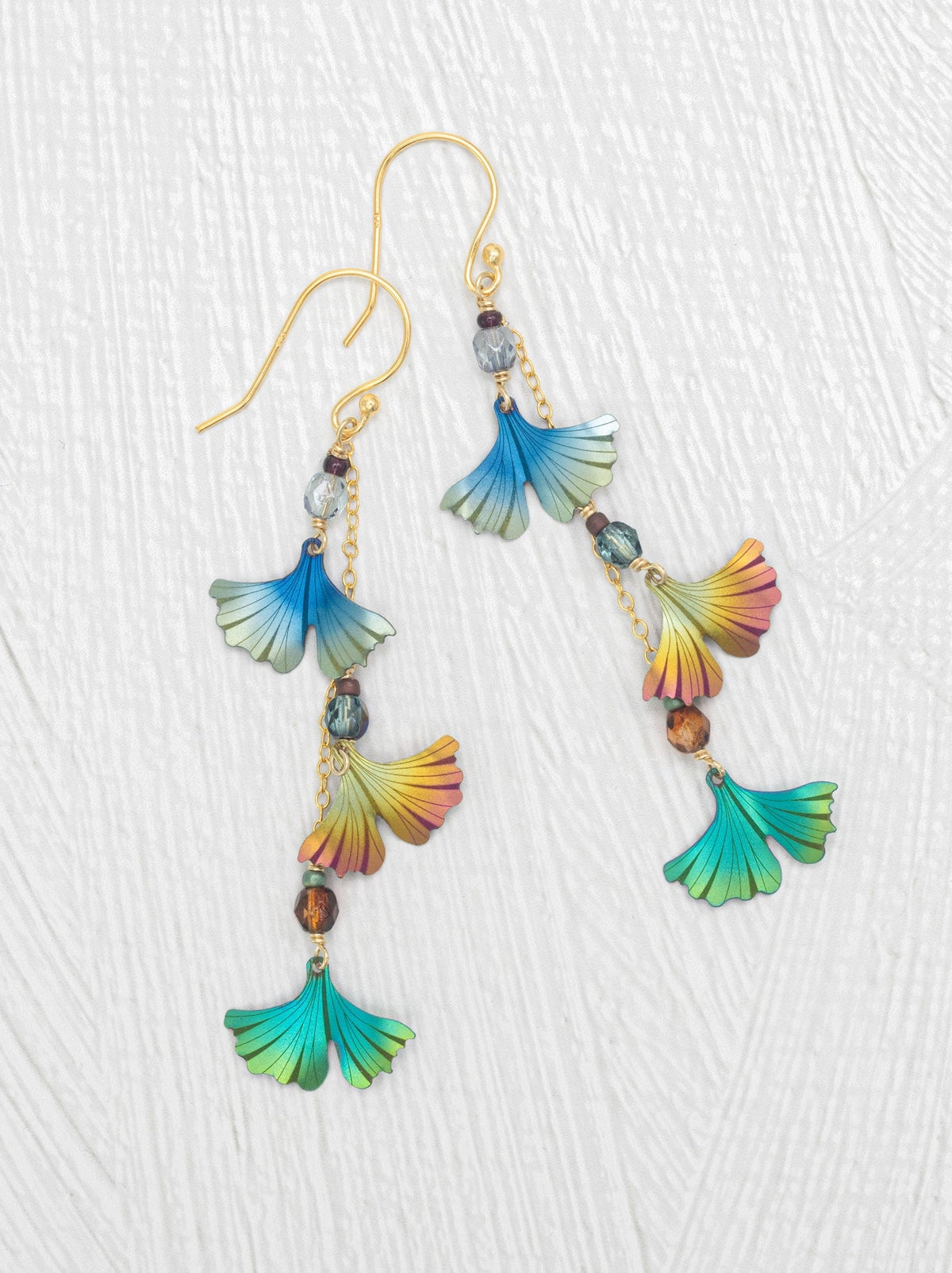 A pair of gold-tone dangle earrings with fan-shaped charms in iridescent blue, green, yellow, and orange, accented by small beads, shown on a textured white background.