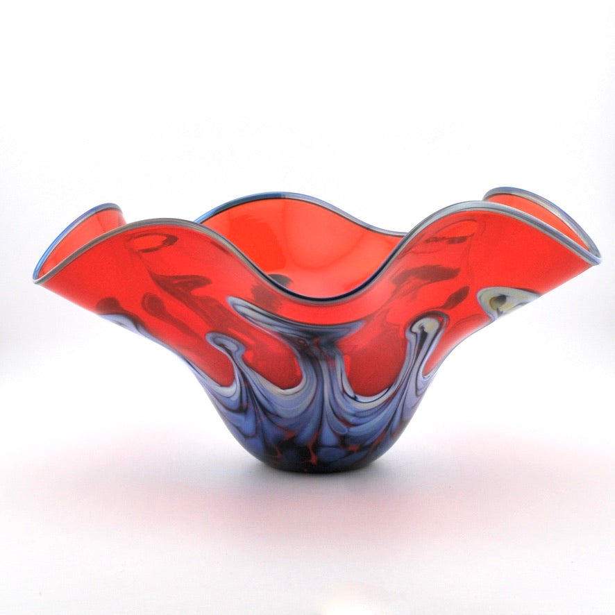 A hand-blown glass bowl with a wavy rim, featuring a vibrant red and blue marbled pattern with white swirls, set against a plain white background.