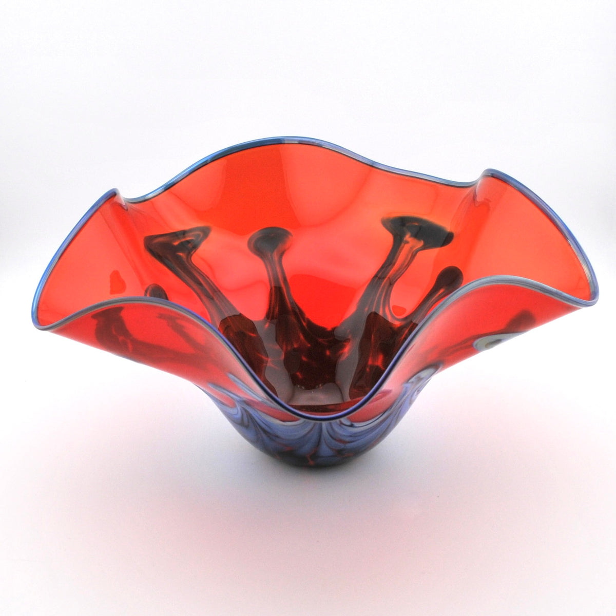 A hand-blown glass bowl with a wavy, flared rim, featuring a vibrant red-orange outer layer and dark purple-black internal swirls, set against a plain white background.