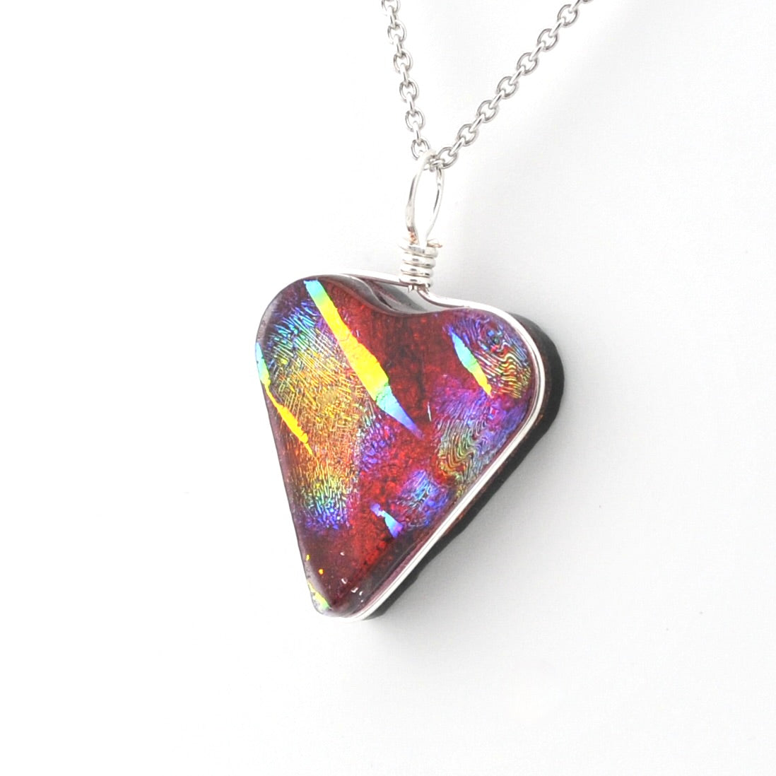 A heart-shaped glass pendant necklace with an iridescent, multicolored pattern of red, purple, blue, and yellow, suspended from a silver chain on a white background.
