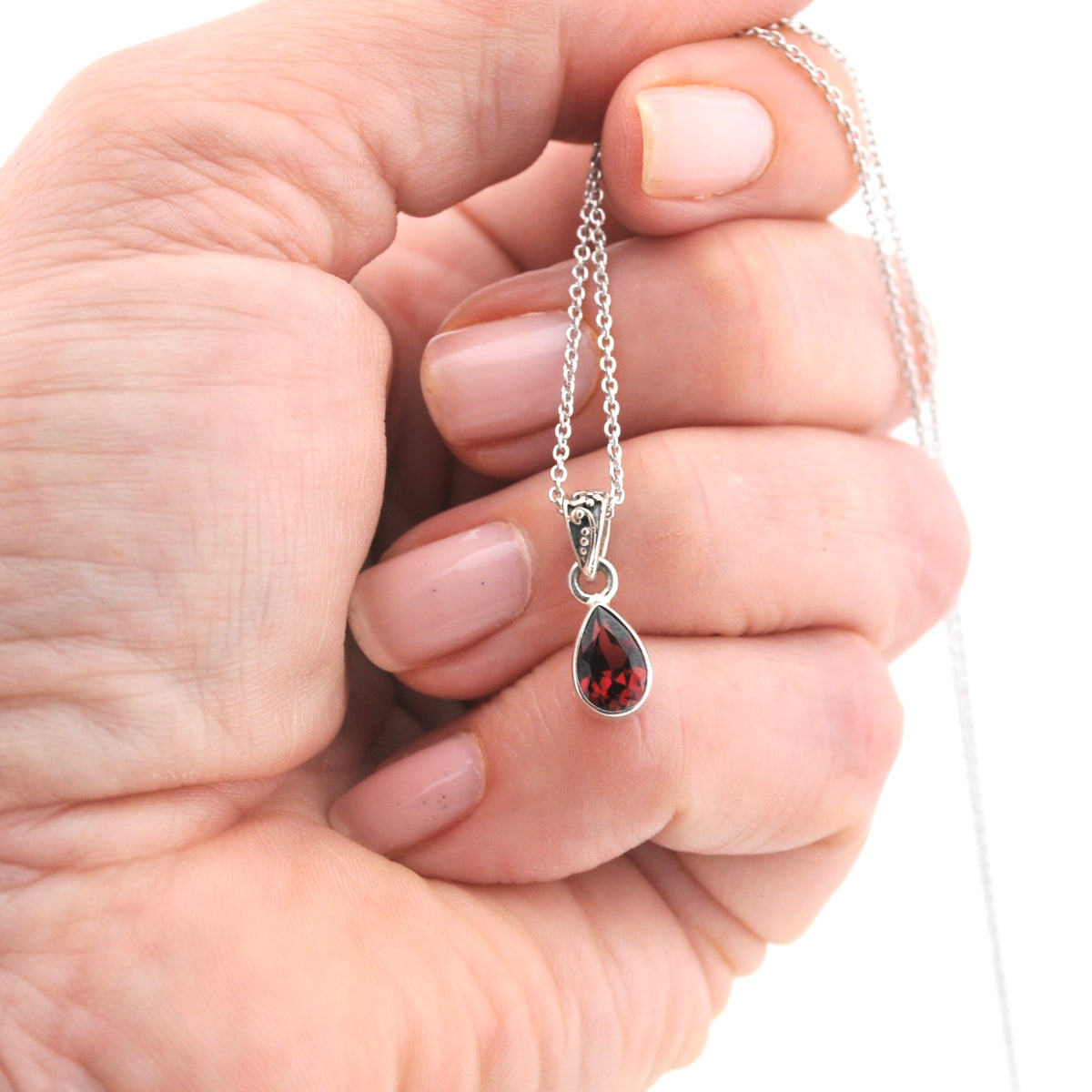 A close-up of a hand holding a silver chain necklace with a teardrop-shaped red gemstone pendant against a white background.