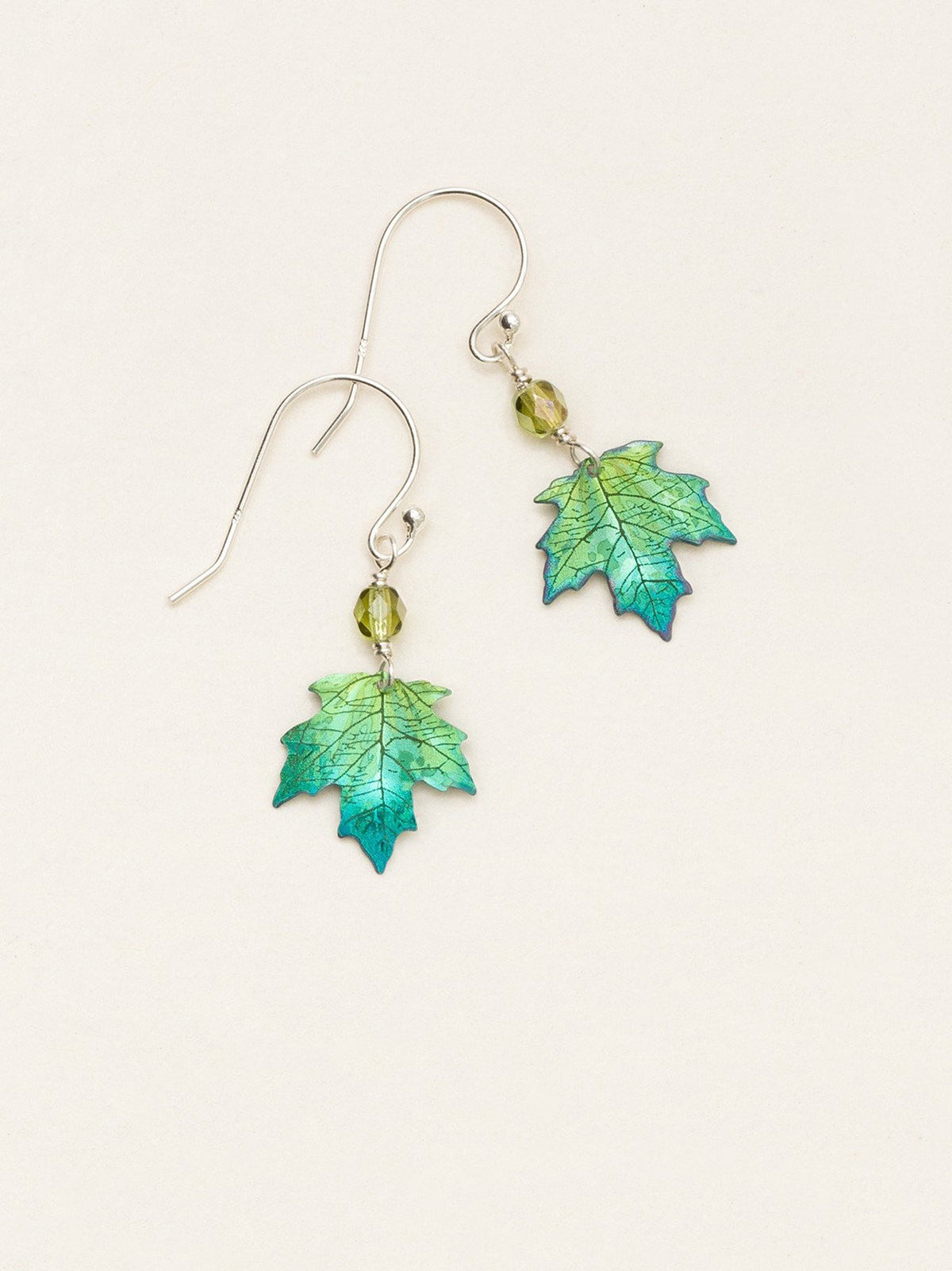 A pair of dangling earrings featuring green and blue iridescent maple leaf charms suspended from silver fishhook wires with small green faceted beads, set against a plain off-white background.